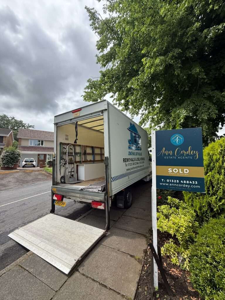 Open-back Moving Van with a Metal Ramp Extended Onto the Pavement Beside a Blue Estate Agents Sign on a Suburban Street, Trees Overhead.